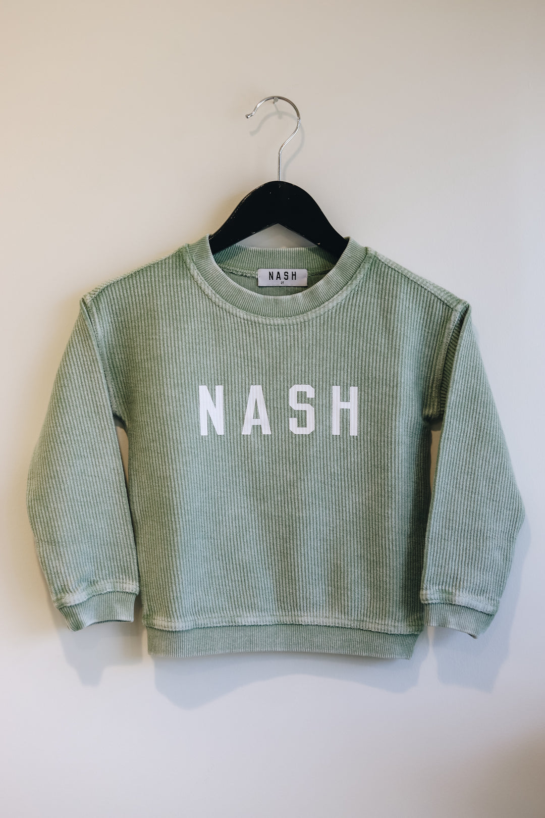Front hanging view of the basil green corded kids crew, with nash screen printed on front